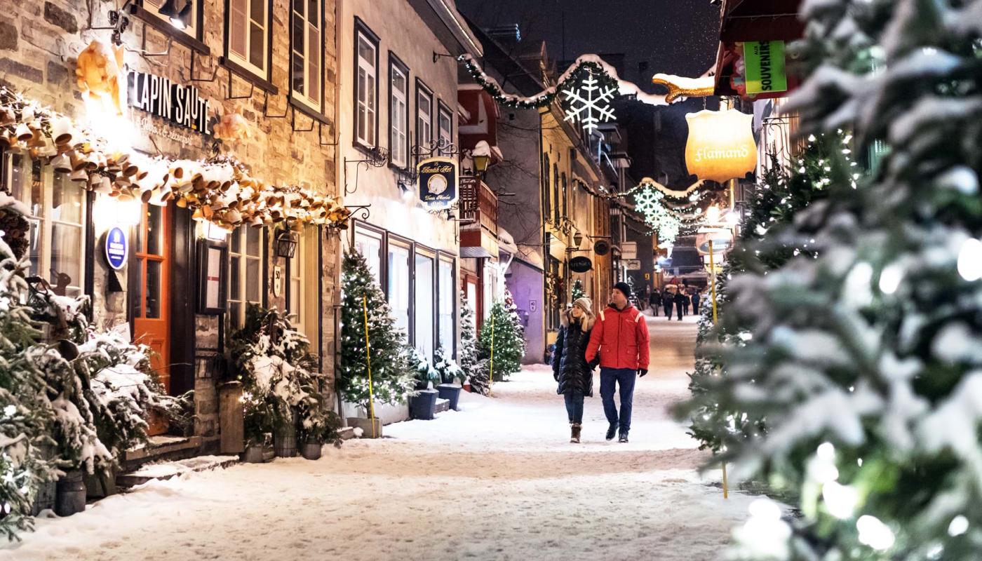 Un couple se promène en soirée durant le temps des fêtes sur la rue du Petit-Champlain enneigée et décorée de sapins illuminés. 