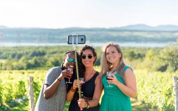 Three smiling girls take pictures of themselves and have a drink in the middle of a vineyard in Ile d'Orleans, in summer.